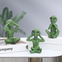 three green alien figurines sitting on top of a white table next to a book