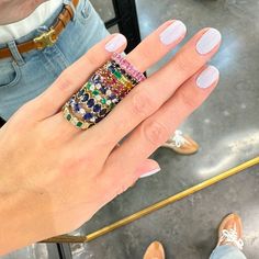 a woman's hand holding a ring with multicolored stones on it and two pairs of shoes in the background