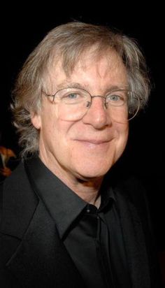 an older man wearing glasses and a black shirt smiles at the camera while standing in front of a dark background