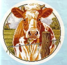 a cross stitch picture of a brown and white cow in a field with a fence