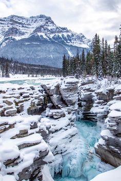 a frozen river surrounded by snow covered rocks and evergreen trees with a mountain in the background