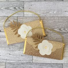 two gold clutches with white flowers on them sitting on top of a wooden floor next to each other