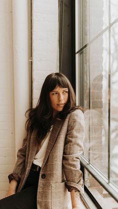 a woman leaning against a window in front of a brick wall wearing a blazer and black pants