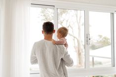 a man holding a baby in his arms while looking out the window at the trees outside