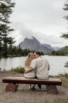 a man and woman sitting on a bench next to a lake with mountains in the background