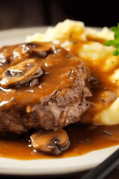 a white plate topped with meat and mushrooms covered in gravy next to mashed potatoes