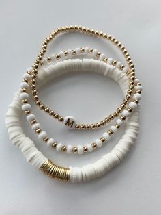 three white and gold beaded bracelets on a white surface with the letter m