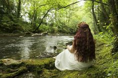 a woman in white dress sitting on mossy ground next to river with trees around her