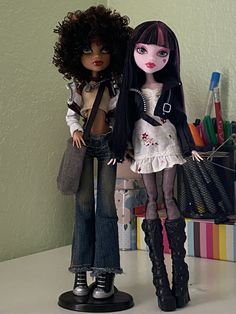 two dolls standing next to each other on a table with pens and pencils in the background