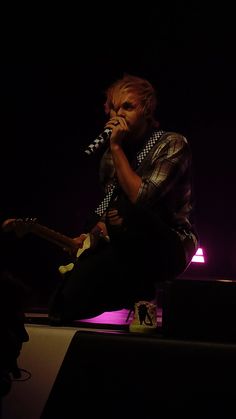 a man sitting on top of a stage while holding a microphone to his mouth and singing into a mic