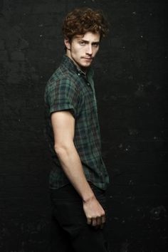 a young man standing in front of a black wall wearing a green shirt and black pants