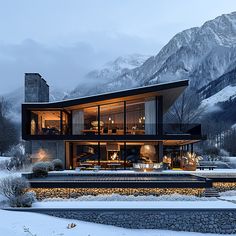 a modern house in the mountains with snow on the ground and lights shining from its windows