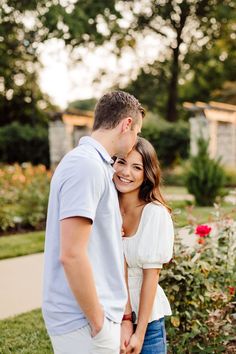 a man and woman standing next to each other in front of some bushes with red flowers