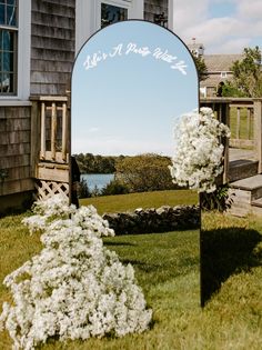 a mirror sitting on top of a grass covered field next to a house with white flowers