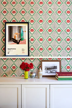 a white dresser topped with a vase filled with red flowers next to a wallpapered wall