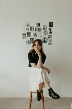 a woman sitting on top of a wooden stool in front of a wall with pictures