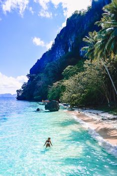 a person swimming in the ocean next to a sandy beach with palm trees and cliffs