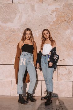 two young women standing next to each other in front of a stone wall wearing jeans and jackets