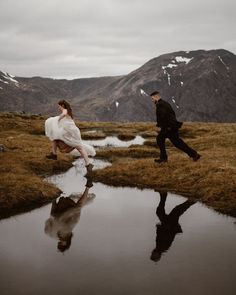 a man and woman walking across a grass covered field next to a small puddle in the ground