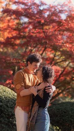 two people standing next to each other in front of trees with red leaves on them