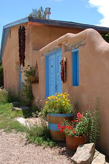 a small adobe house with blue doors and flowers in the front yard, on a sunny day