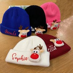 four knitted hats with reindeer heads and name written on the caps are sitting on a wooden surface