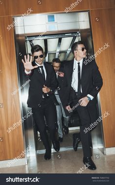 two men in suits exiting an elevator with their hands up to the door and one man wearing