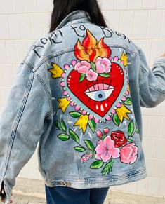 a woman wearing a jean jacket with an embroidered heart and flowers on the back, standing in front of a white brick wall