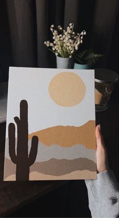 a person holding up a piece of art with a cactus in the foreground and a potted plant next to it