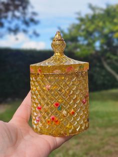 a hand holding a gold colored glass jar with red, green and yellow decorations on it