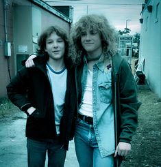 two young men standing next to each other in front of a brick wall and building