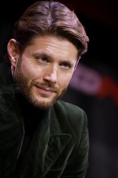 a man with long hair and beard wearing a green jacket looks at the camera while sitting down