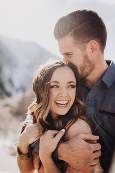 a man holding a woman in his arms and smiling at the camera with mountains in the background