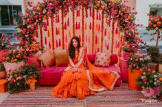 a woman in an orange dress sitting on a pink couch surrounded by flowers and greenery