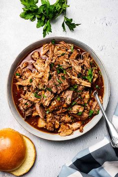 a bowl filled with meat and garnish next to bread on a white surface