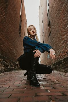 a woman sitting on the ground in an alleyway with her legs crossed and wearing black boots