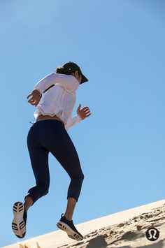 a woman in white shirt and black cap running on sand dunes with blue sky behind her