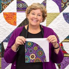 a woman holding up a piece of paper in front of a quilted wall hanging