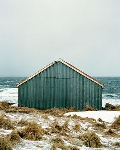 an old green building sitting on top of snow covered ground next to the ocean with waves coming in