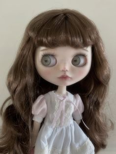 a doll with long brown hair wearing a white dress and blue eyes is standing in front of a wall