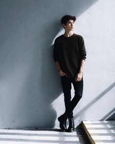 a young man leaning against a wall with his hands in his pockets and shoes on