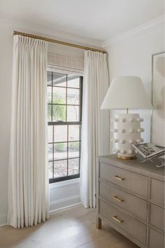 a bedroom with white curtains and a lamp on top of a dresser next to a window