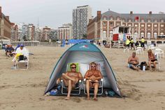 two men sitting in chairs on the beach next to a blue tent and some buildings