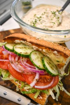 a sandwich with cucumbers, tomatoes and onions on it next to a bowl of dressing