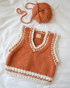 an orange and white knitted vest laying on top of a bed next to a ball of yarn