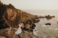 a bride and groom standing on the rocks by the ocean