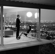 a man and woman standing in front of a large window overlooking the city at night