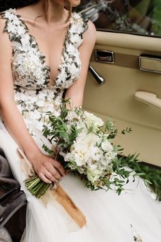 a woman in a wedding dress sitting on the back of a car holding a bouquet