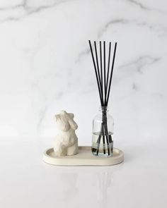 a white marbled counter top with a glass vase filled with reeds and a bear figurine next to it