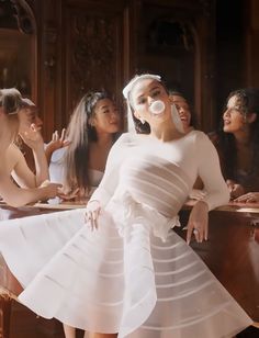 a woman in a white dress standing next to other women sitting at a table and blowing bubbles